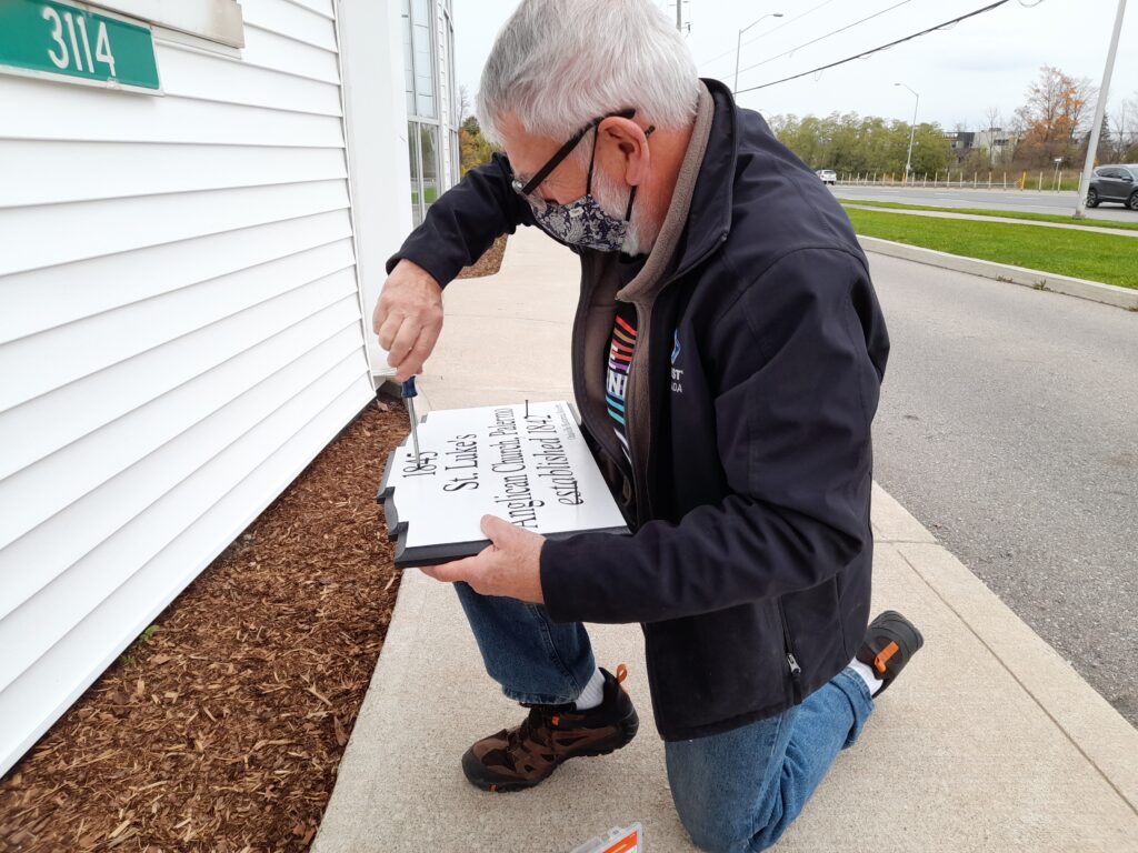 George Chisholm inserts screws into the new plaque, preparing it to be installed on the exterior wall of St. Luke's church. He is kneeling on the sidewalk in front of the white vinyl siding of the church.